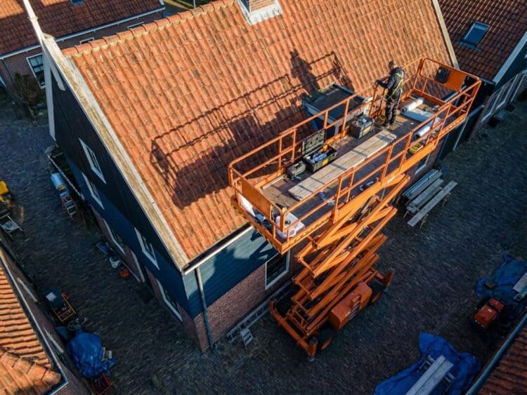 Oranje hoogwerker bij rood pannendak van Nederlands huis, hydraulische mechanismen en veiligheidsrailing zichtbaar