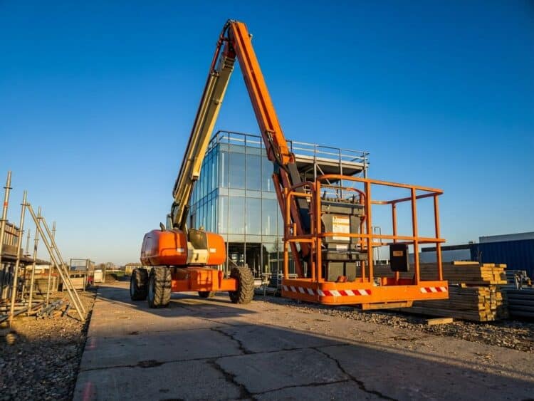 Oranje hoogwerker met uitgeschoven arm tegen blauwe hemel op bouwplaats met steigers en betonnen ondergrond