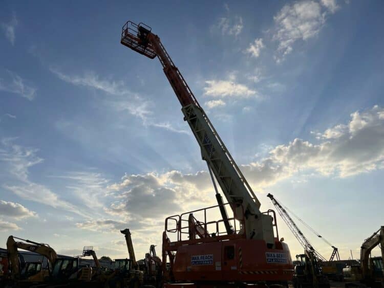 Oranje hoogwerker steekt dramatisch omhoog tegen blauwe hemel met witte wolken, constructieapparatuur aan de basis