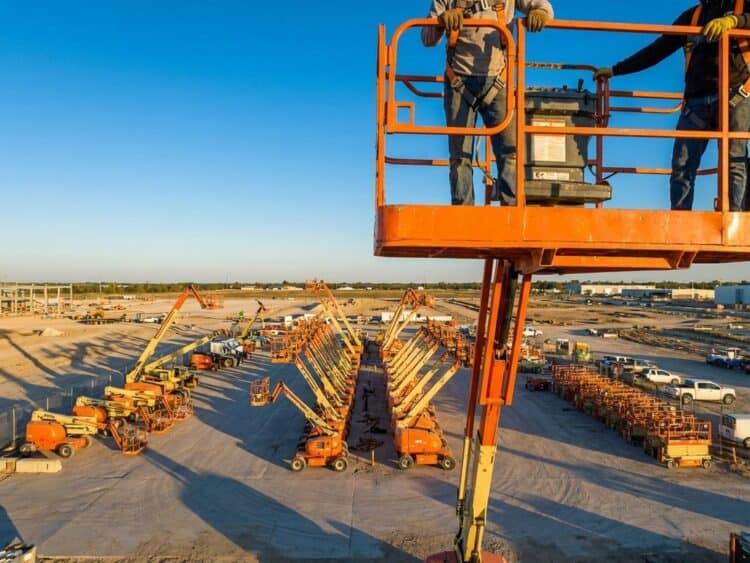 Oranje hoogwerker op bouwplaats met arbeiders op platform, gele en oranje luchtplatforms op georganiseerde werkplaats