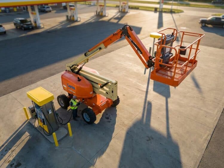 Oranje hoogwerker bij geel tankstation tijdens gouden uur, hydraulische details en veiligheidsreling zichtbaar