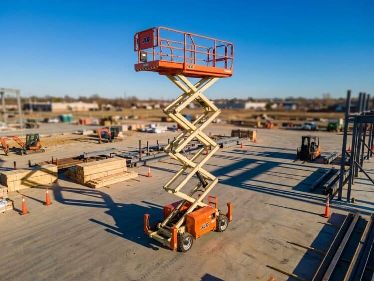 Oranje hoogwerker met uitgeschoven platform op bouwplaats tegen blauwe hemel, omringd door bouwmaterialen