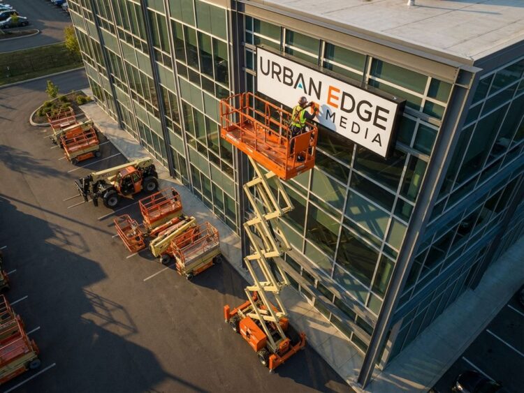 Oranje schaarlift met werknemer die verlichte reclamebord installeert op commercieel gebouw, verhuurmachines zichtbaar