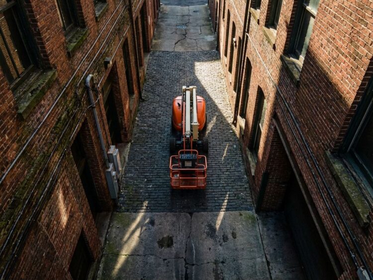 Compacte oranje schaarlift navigeert door smalle stedelijke steeg tussen hoge bakstenen gebouwen
