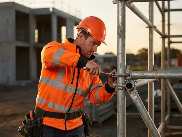 Bouwvakker in oranje veiligheidskleding past metalen steigerbuizen aan op bouwplaats tijdens gouden uur