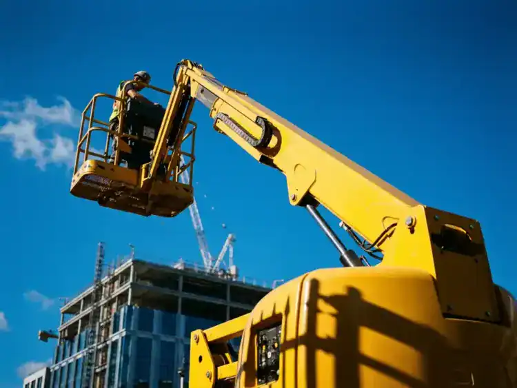 Gele hoogwerker van 12 meter volledig uitgeschoven met bouwvakker in mand op commerciële bouwplaats tegen blauwe lucht