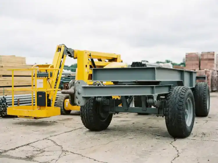 Zware aanhangwagen met stalen frame achter gele hoogwerker op betonnen ondergrond, bouwplaats met materialen op achtergrond