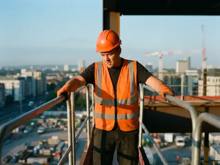 Bouwvakker in oranje veiligheidsvest staat op smalle stalen balk hoog boven bouwplaats, grijpt leuningen vast