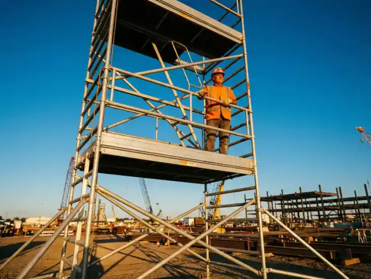 Bouwvakker in gele veiligheidsvest controleert leuningen op hoge mobiele steiger tegen blauwe hemel op bouwplaats