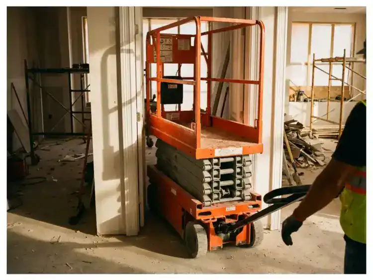 Oranje schaarlift met verhoogd platform voor witte deuropening op bouwplaats met betonnen vloer