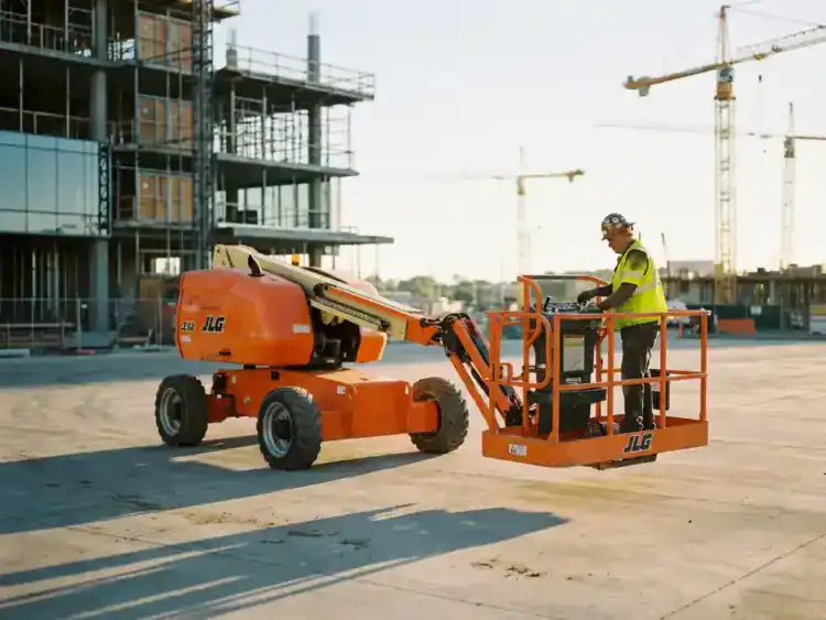 Oranje schaarlift met werknemer in veiligheidsvest op bouwplaats, kranen op achtergrond in ochtendlicht