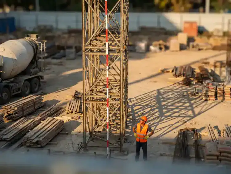 Hoge metalen steiger naast rood-witte hoogtemeter met bouwvakker in oranje veiligheidsvest op drukke bouwplaats