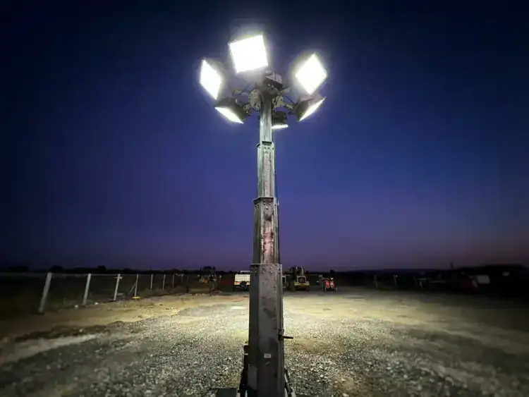 Industriële lichtmast volledig uitgeschoven op bouwterrein bij schemering, felle schijnwerpers verlichten grindbodem vanuit laag camerastandpunt.