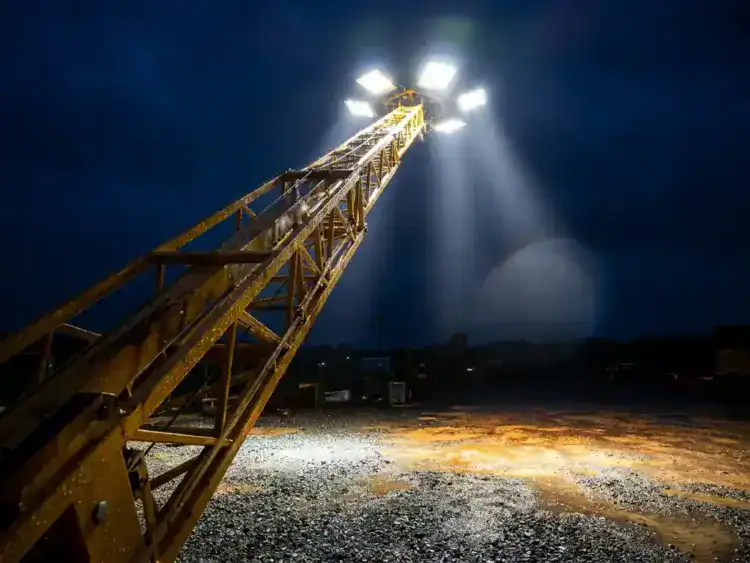Verlichte mobiele lichtmast op nachtelijke bouwplaats, krachtige schijnwerpers verlichten nat grind, geel stalen frame tegen donkerblauwe lucht.
