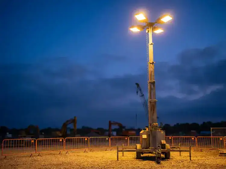 Uitgeschoven mobiele lichtmast op een bouwplaats bij schemering, verlicht grind met veiligheidshekken en zwaar materieel op de achtergrond.