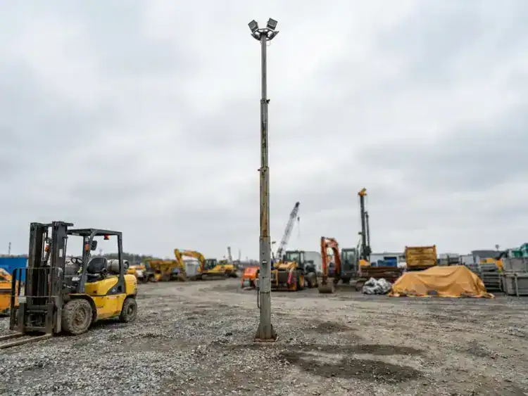 12 meter hoge stalen lichtmast op bouwterrein, met gele heftruck ernaast voor schaalvergelijking op grindondergrond.