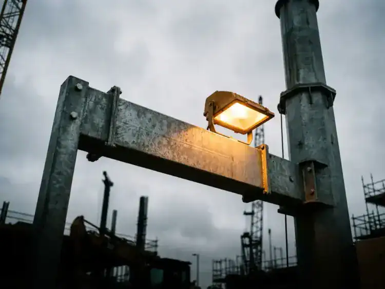Stalen uitleggerarm van een industriële lichtmast, van onderen gefotografeerd tegen bewolkte bouwplaatslucht met warm amber schijnwerperlicht.